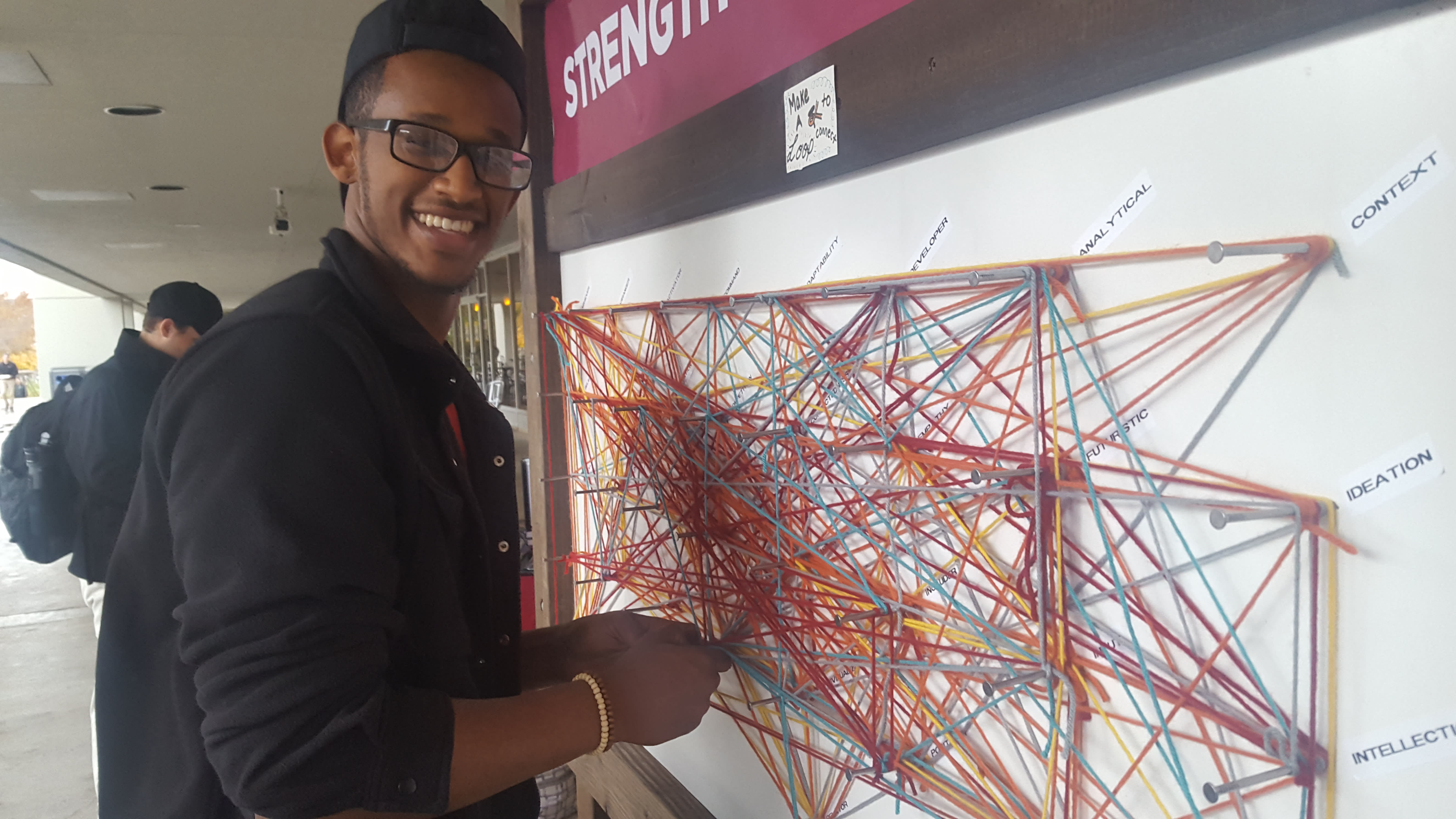 A student participates in a strengths pop up event. He is wrapping colored yarn around his top 5 strengths as a part of a larger web representing community members' top 5 strengths.