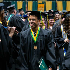 A smiling culinary student in a black graduation cap and gown walks through a crowd of fellow graduates at a University of Alaska Anchorage commencement ceremony. He wears a green and gold honor cord and medal while holding his diploma, surrounded by joyful applause