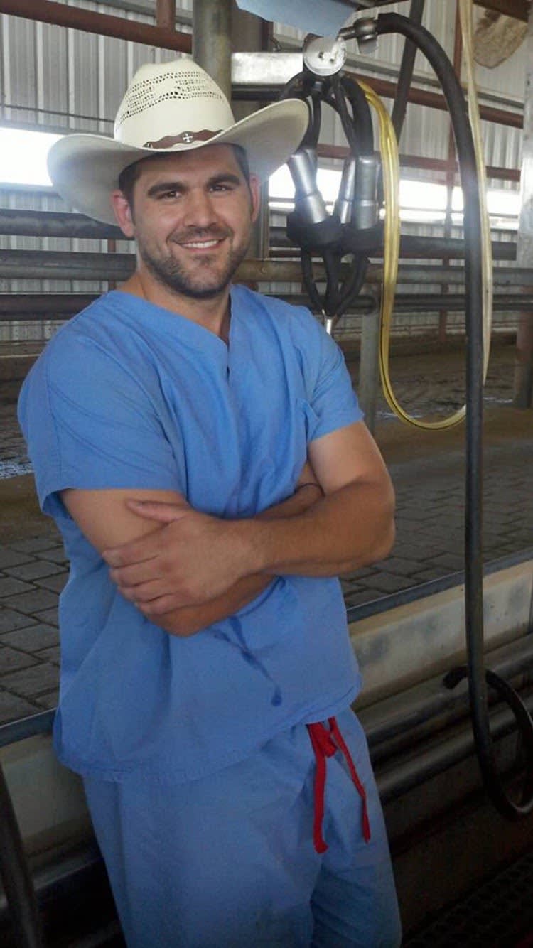 Austin Ayars wearing blue, medical scrubs and a white cowboy hat. He is leaning against farm equipment for cows