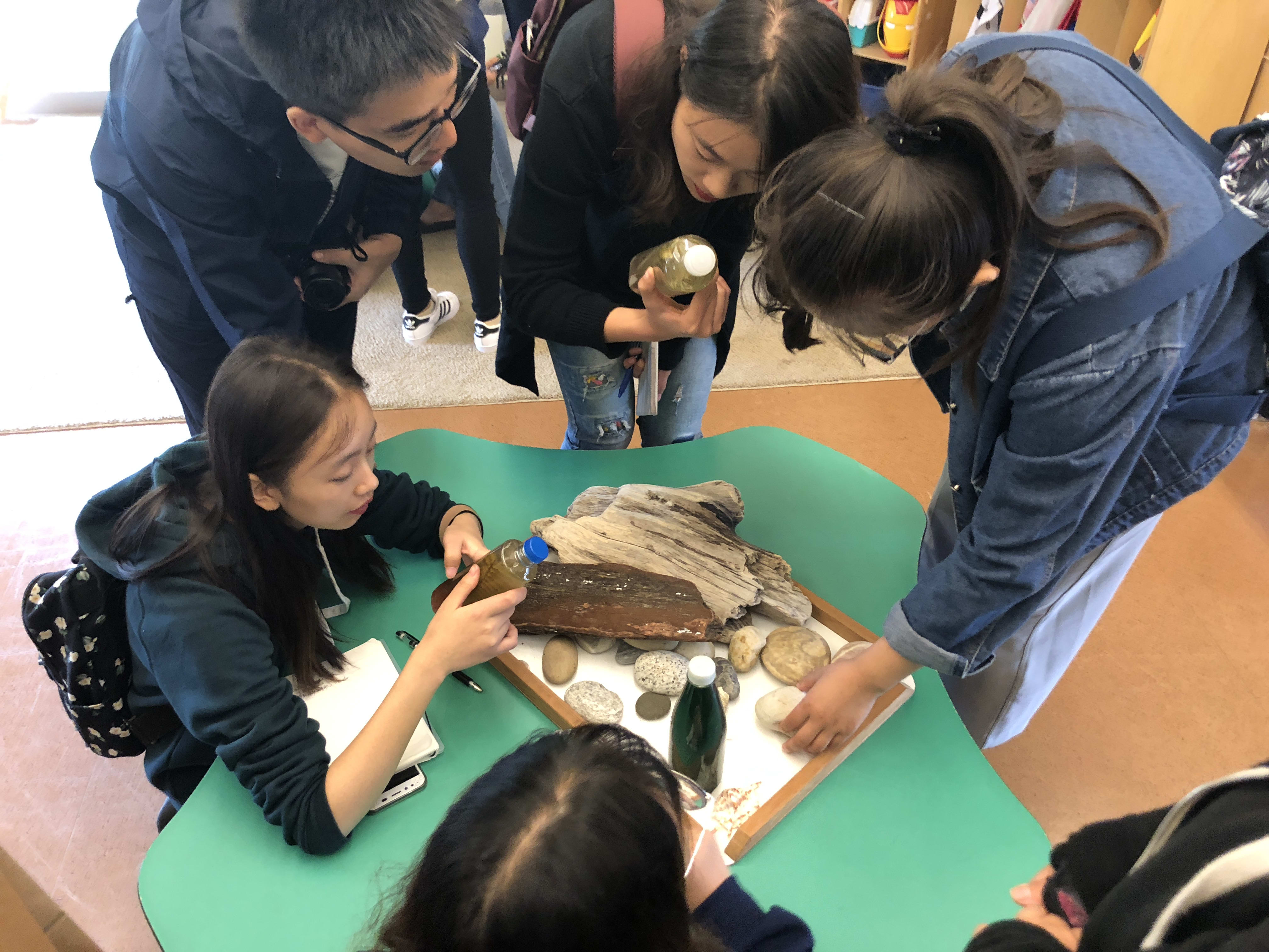 A group of Chinese students at a table investigating the wood, rocks, and bottles of dirty water on the table.