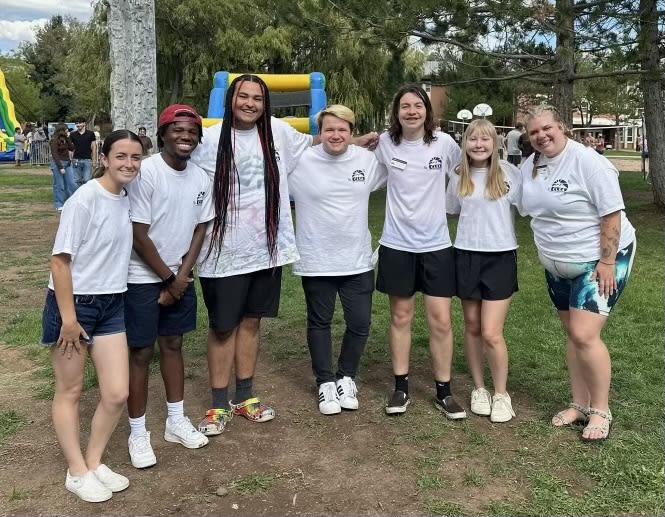 6 CLSA executives and 2 advisors lined up shoulder to shoulder wearing white branded t-shirts on a half grassy and dirt field. In the background to the left are more students at the event and inflatables and a tall rock wall.
