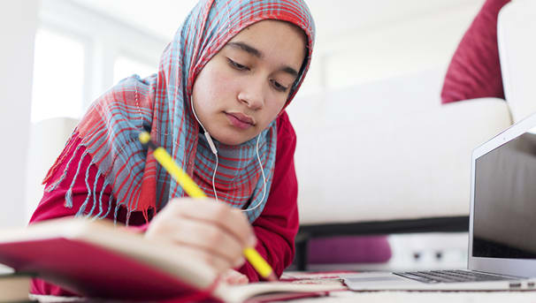 A young student taking notes in a small journal. She sits on the floor, next to an open laptop
