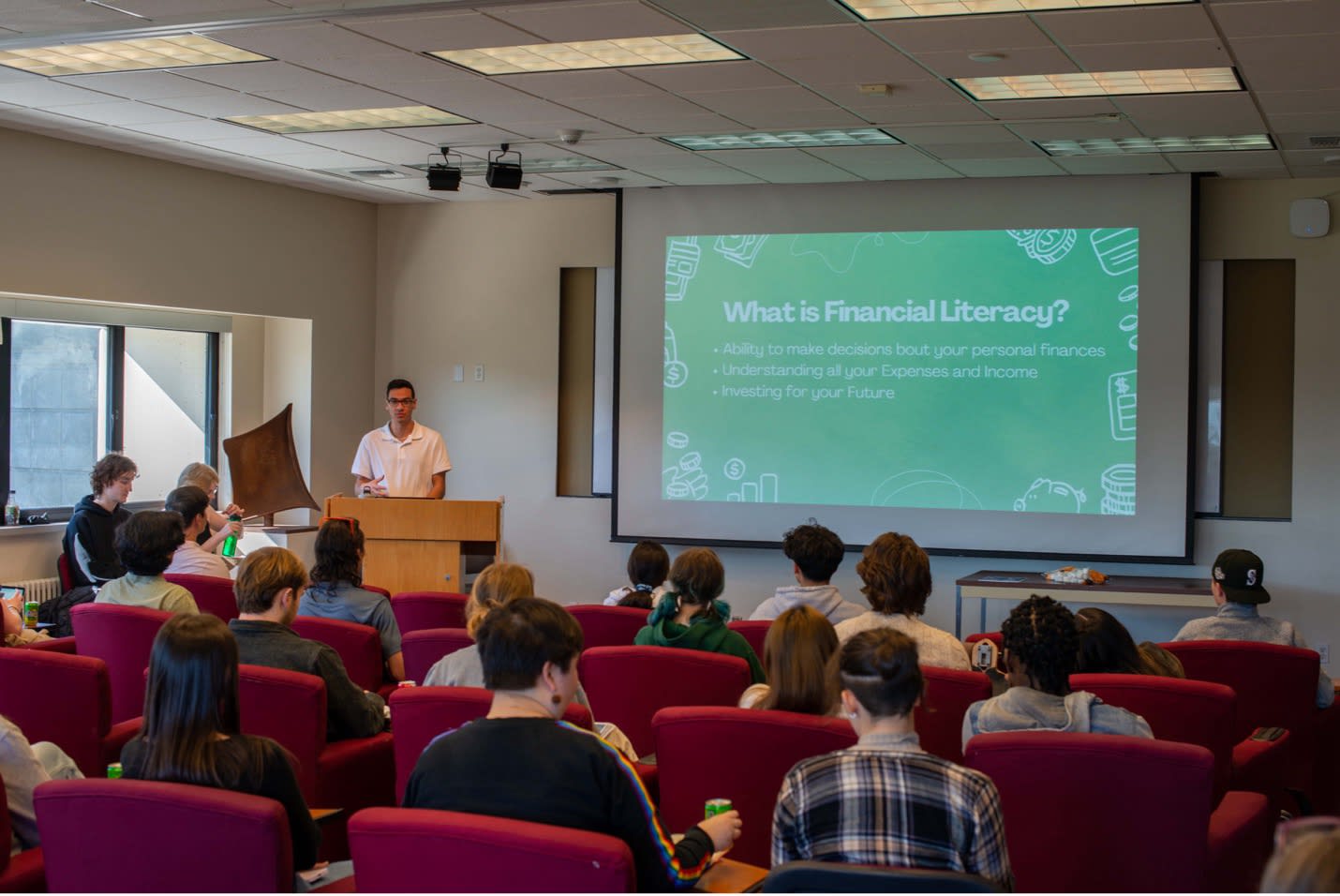 A person stands behind a podium at the front of a room with a slide on screen reading "What is Financial Literacy?"