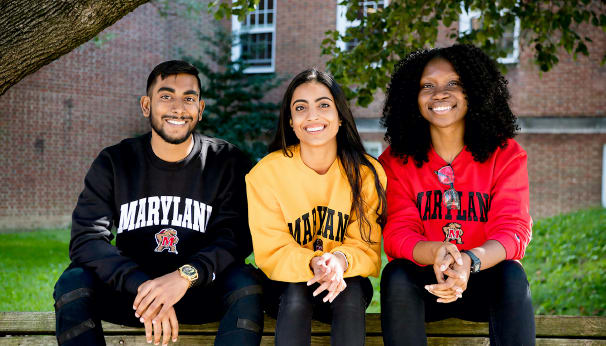 Three UMD students--a man and two women--sitting outside and wearing Maryland-themed sweatshirts.