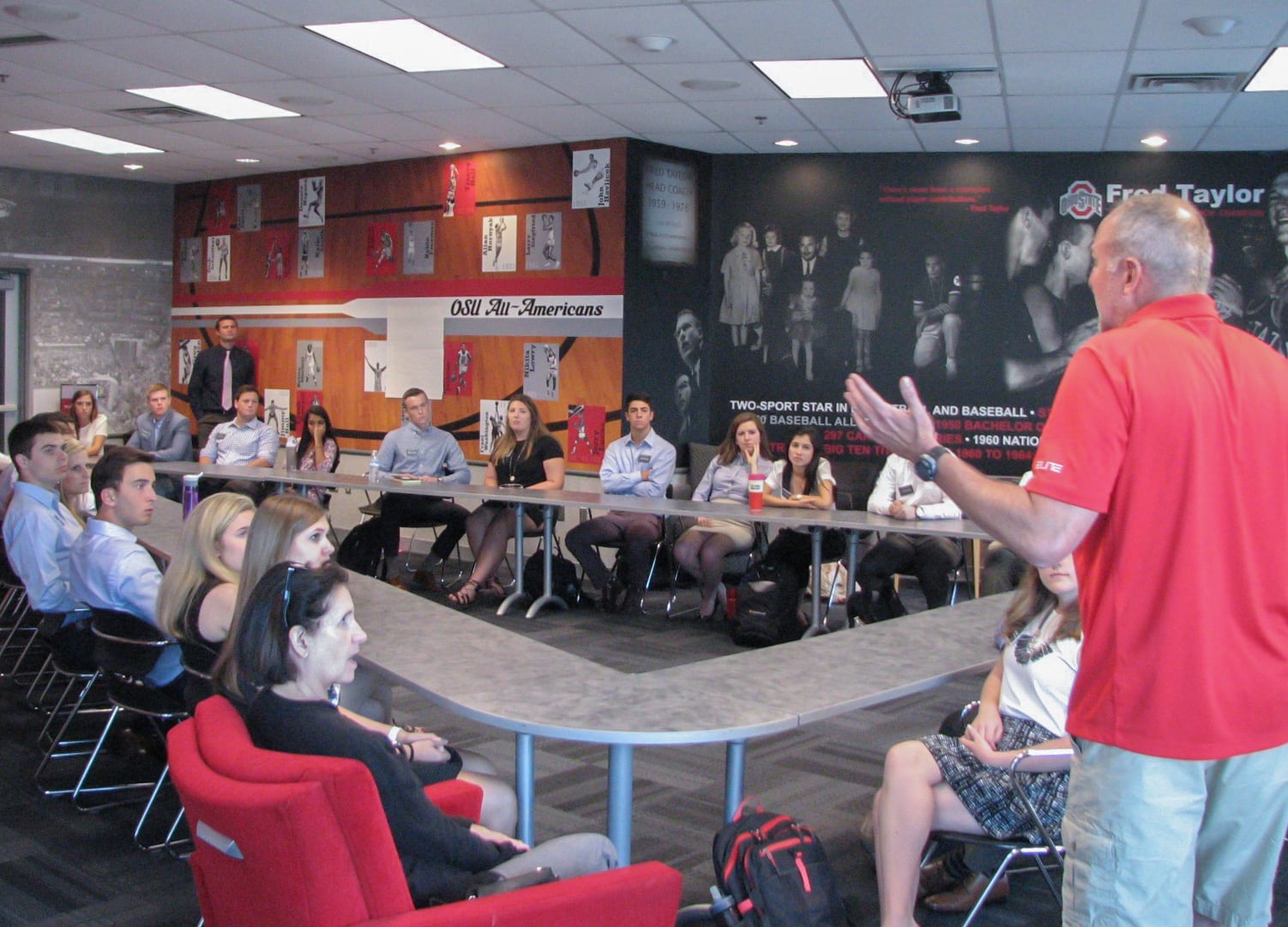 A professor teaches students who are sitting around tables in a classroom