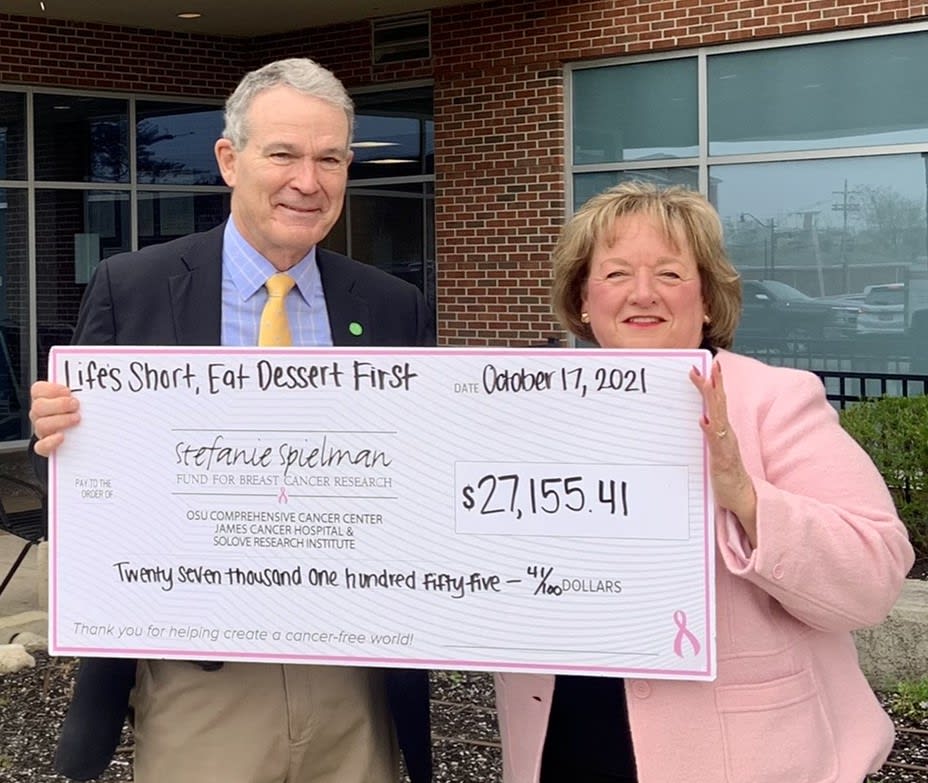 Two people hold a check for $27,155.41 in front of an office building 