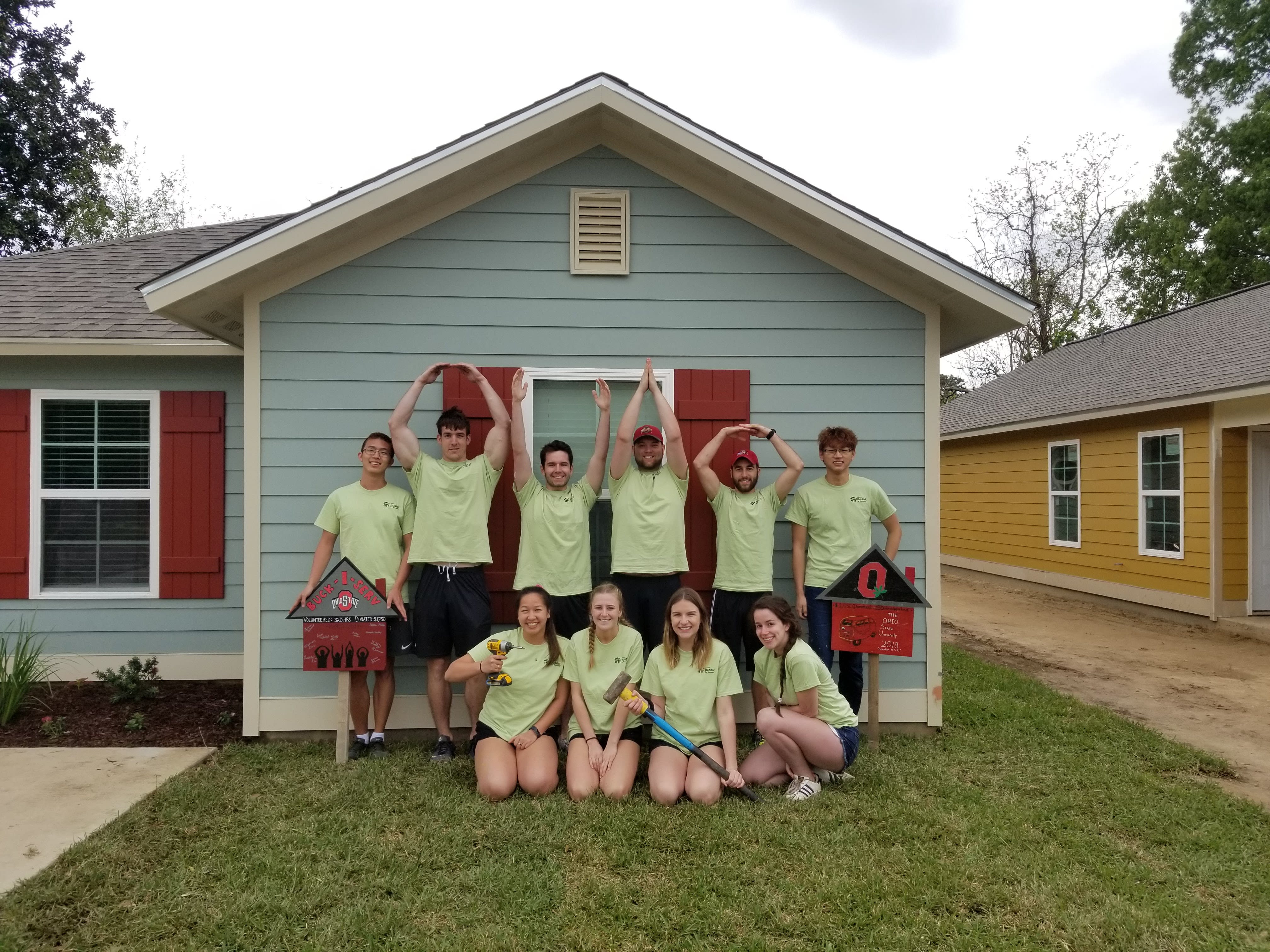 Volunteers with various tools pose before a colored house. The ones with free hands do O-H-I-O with their arms