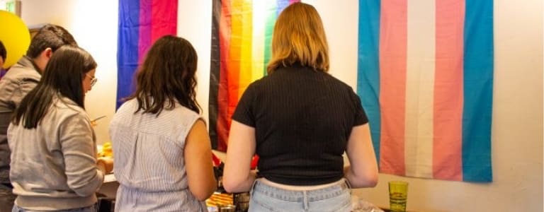 4 students grabbing food from a table. 3 flags representing LBGTQ+ on the wall behind.