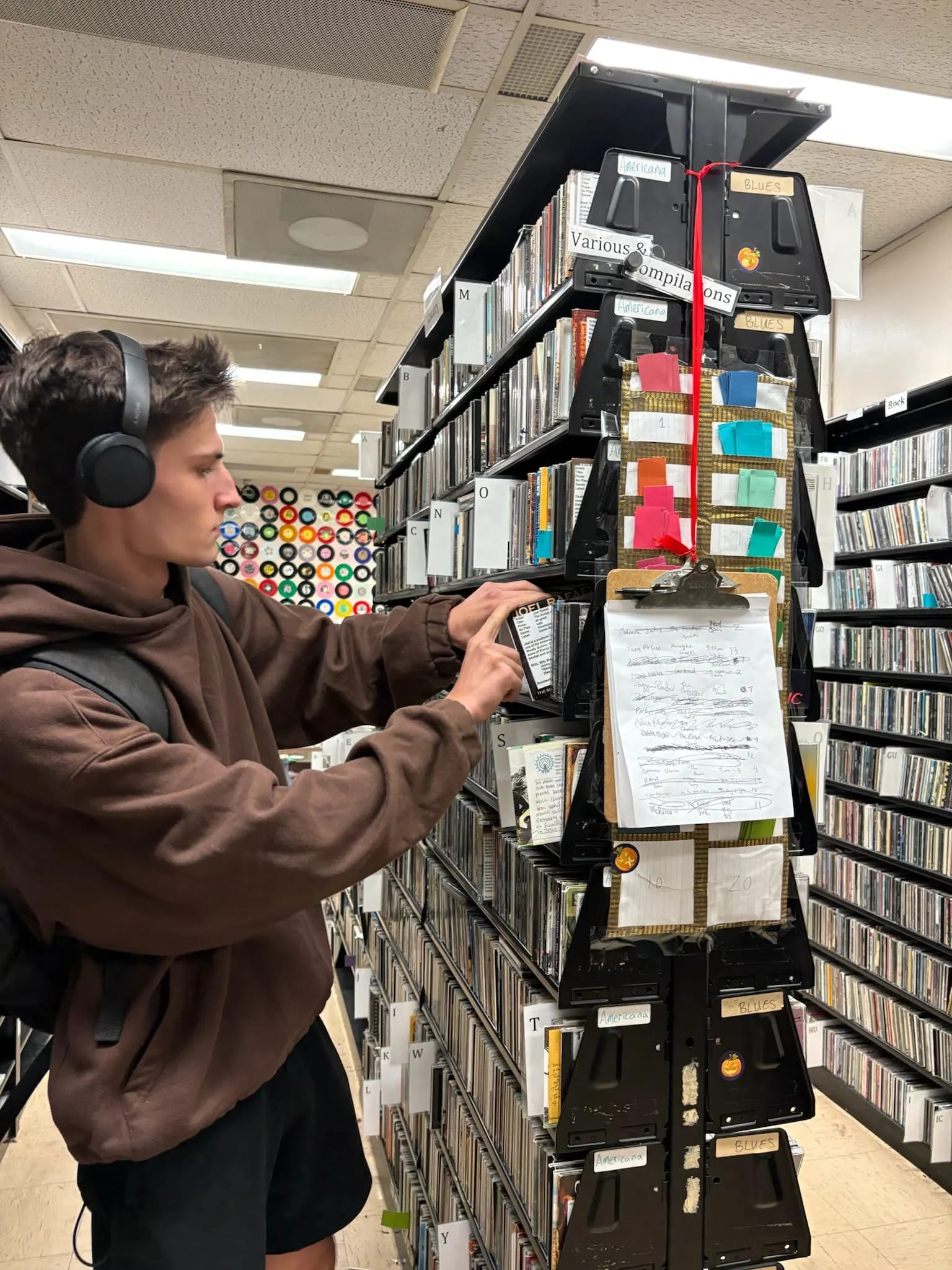 Person wearing headphones browses CDs in a music library aisle lined with shelves and labeled sections.