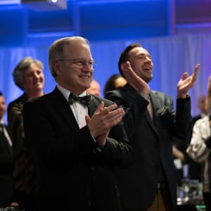 Attendees at the 2024 Celebrity Chefs Invitational give a standing ovation. A man in a bow tie and glasses claps enthusiastically in the foreground, with other formally dressed guests smiling and applauding in the background.