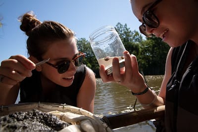 Two students examine the specimen within a glass jar. One is holding a net full of river content beneath their arm.
