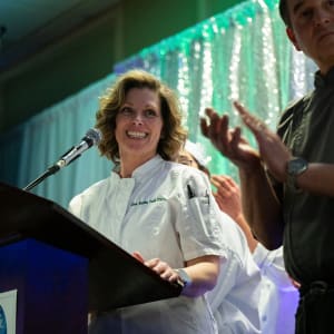 Chef Puff in a white coat smiles while speaking at a podium during the 2024 Celebrity Chefs Invitational. Behind her, fellow chefs and attendees applaud under sparkling green and silver stage lights