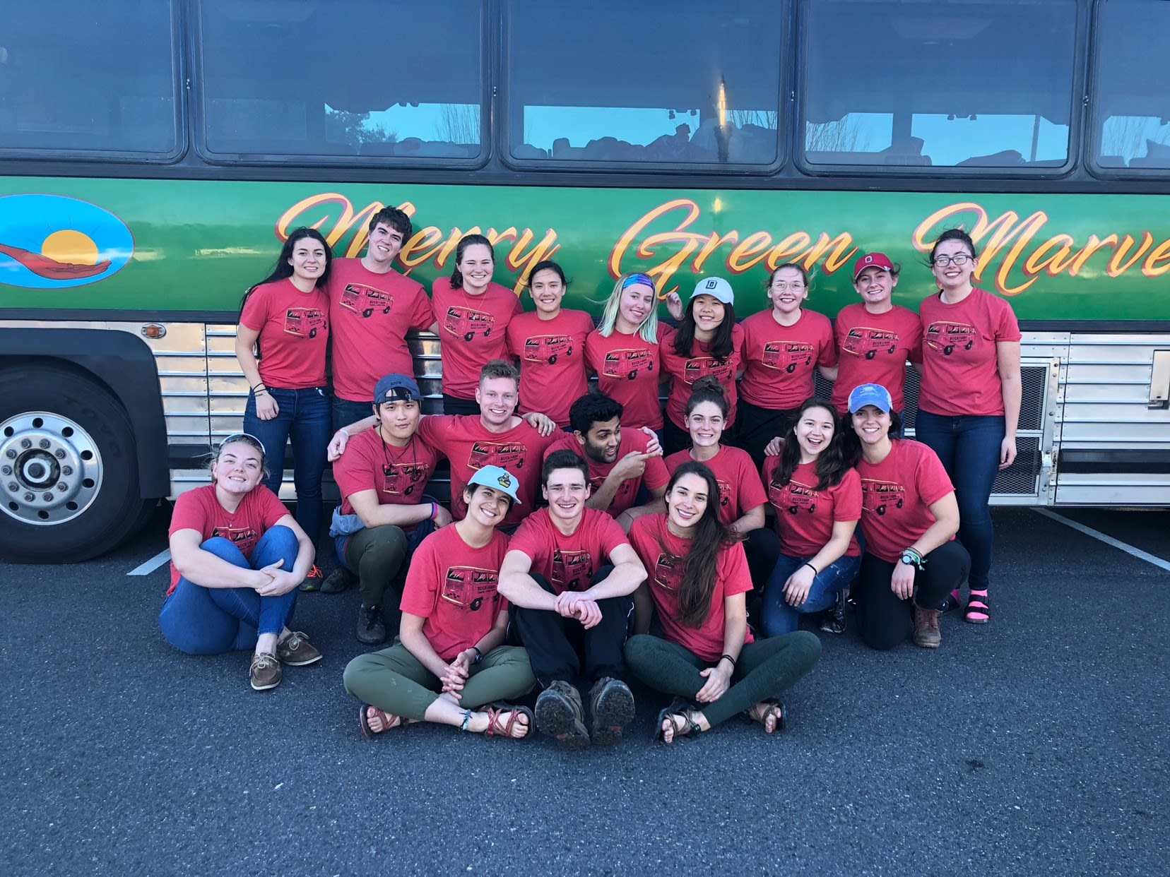 Volunteers in red t-shirts smile before a large bus. On a green background reads 'Merry Green Marvels' on the bus