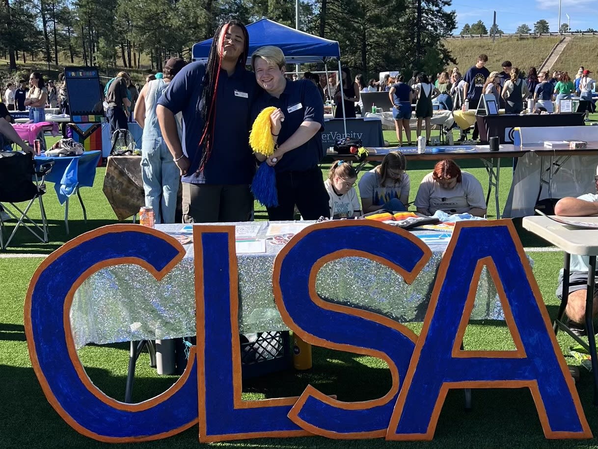 Large wooden blue and orange letters that spell CLSA are in the center. Behind them is a table with a sparkling table cloth and 2 CLSA executives posing. Near them and in the background is other student clubs and organizations tabling. Some people are sitting and standing in the background.