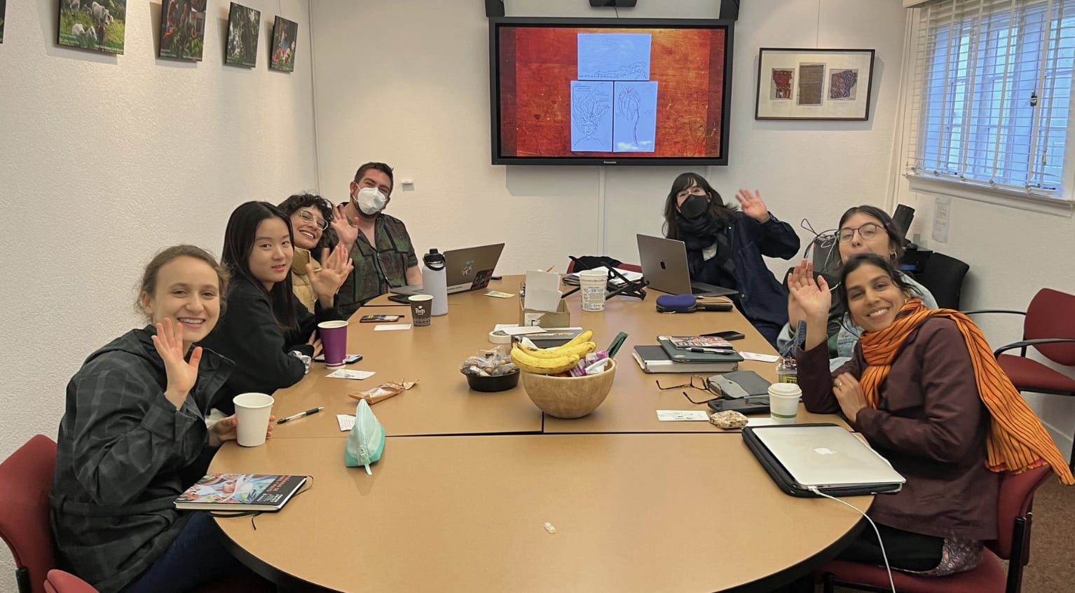 Students sit around a seminar table and wave at the camera, in front of a screen with images of white paper on it. 