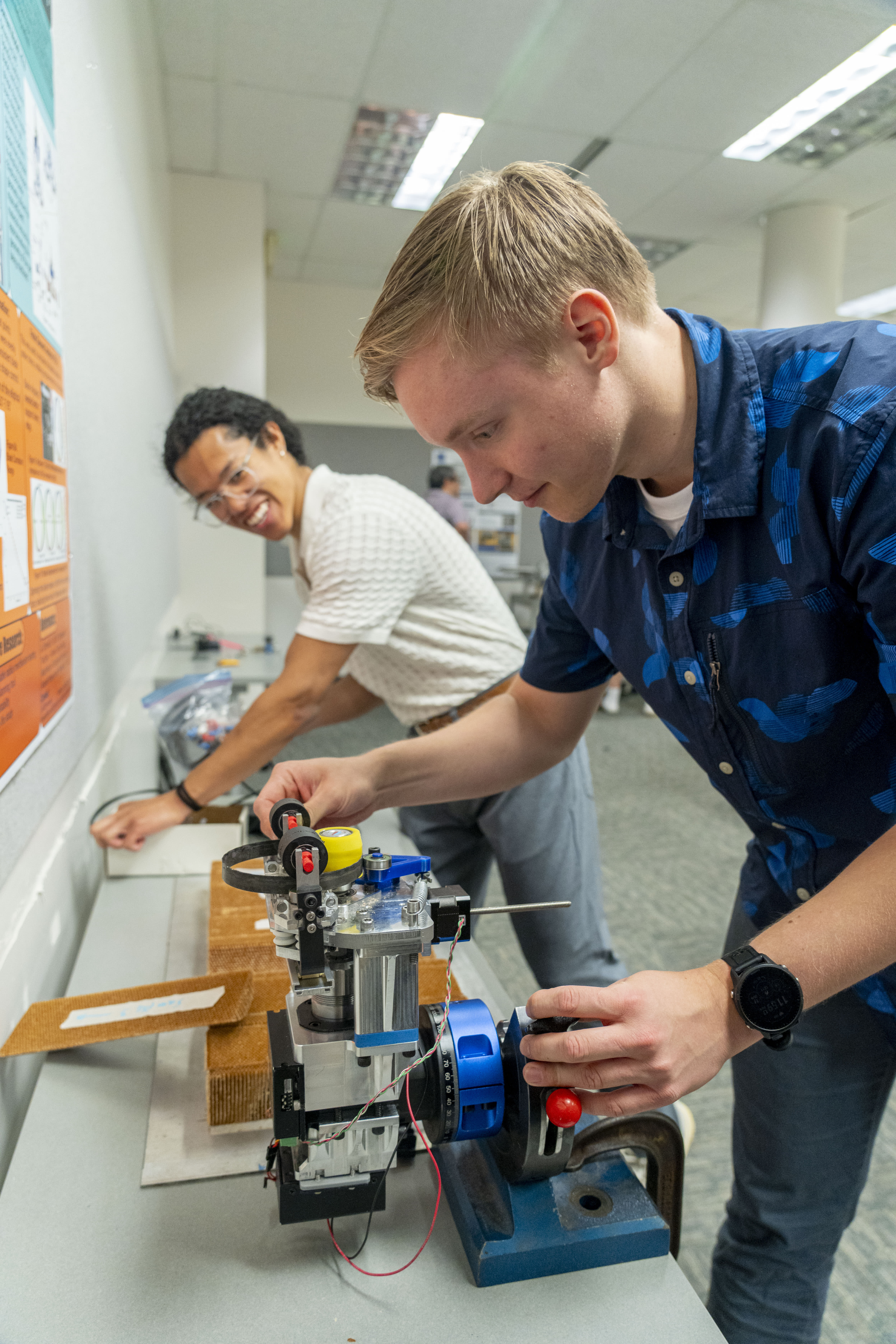 Two men work on a robotic device. One adjusts a component, the other smiles, looking at a poster.