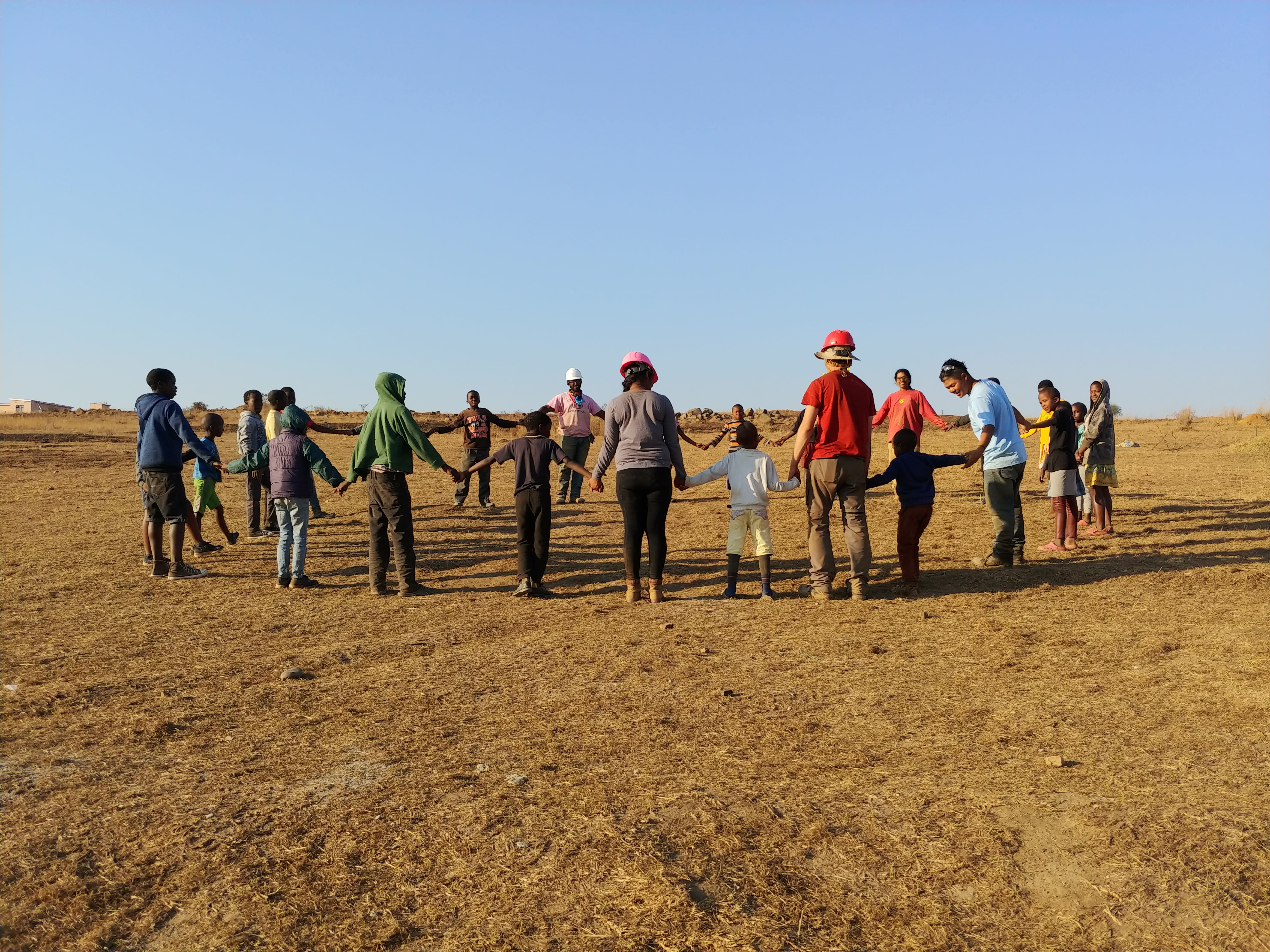 End-of-day work circle where travel team, community members, and children are all holding hands