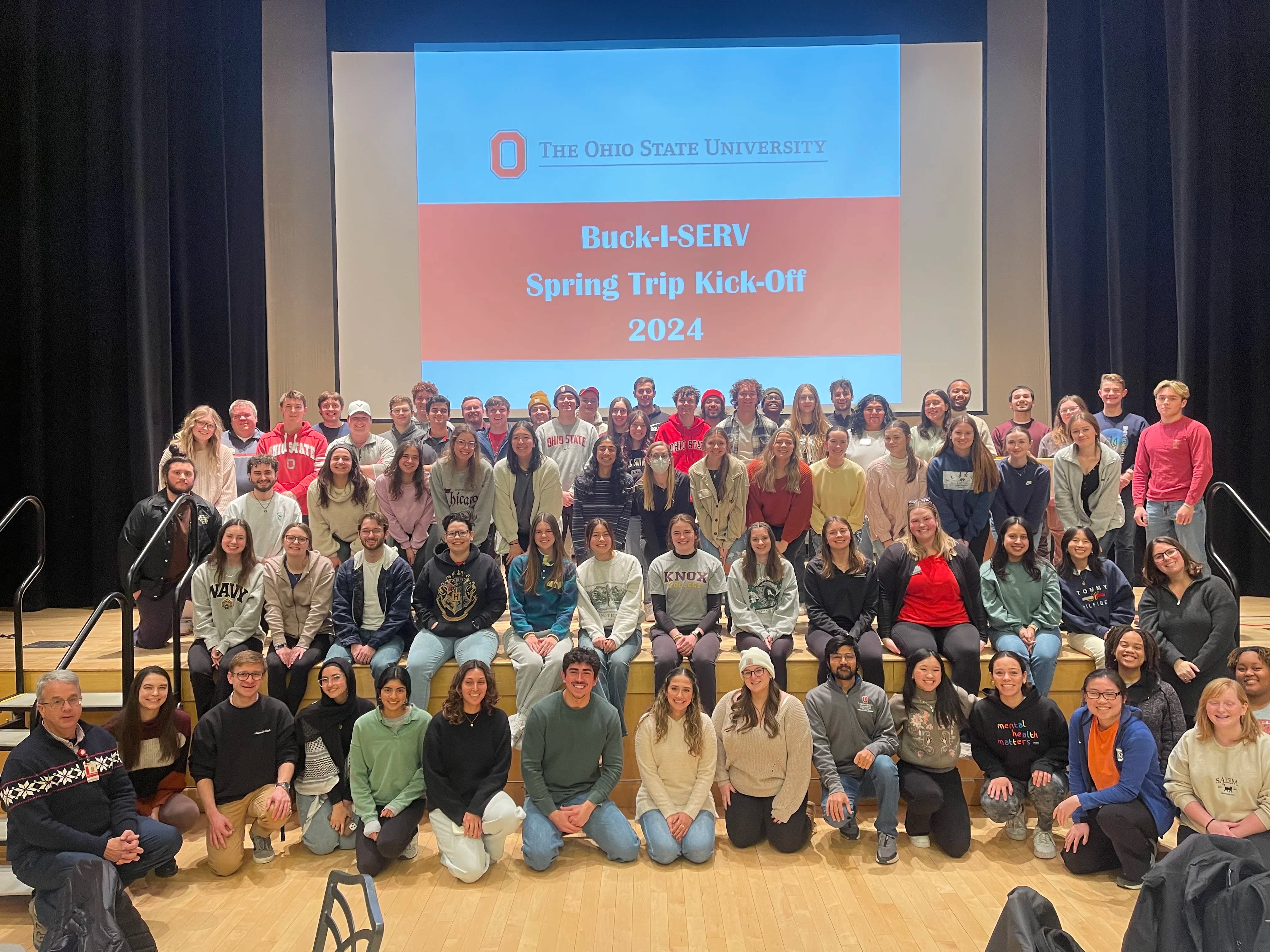 A large group of students posed in front of the projection 'Spring Trip Kick-Off 2024' slideshow 