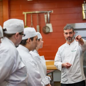 A chef instructor in a white coat gestures while teaching a group of culinary students in a professional kitchen. The students, dressed in white uniforms and hats, attentively listen as they stand in front of a red brick wall with hanging kitchen tools.