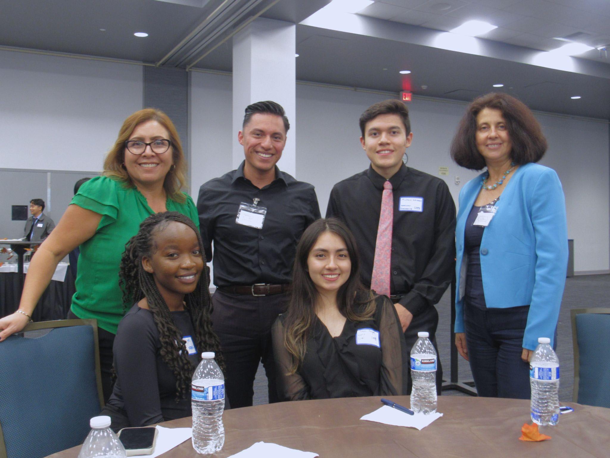 Six people in professional dress and wearing nametags sit and stand around a table in a large conference room.