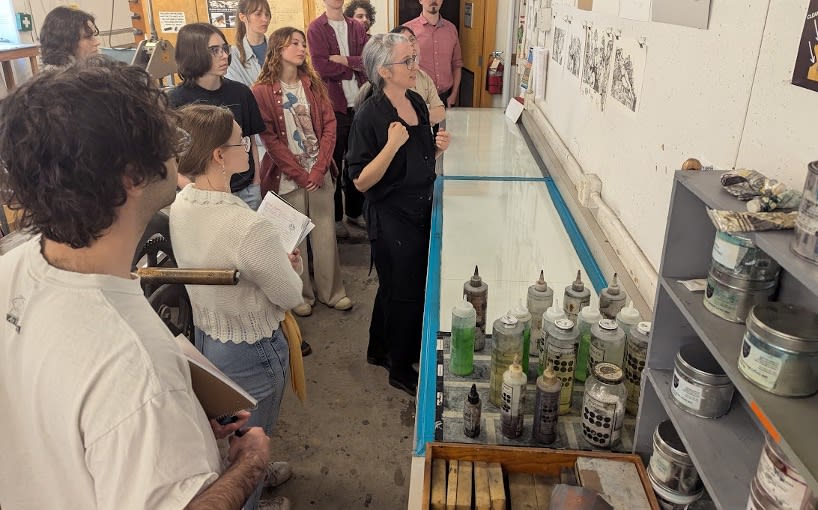 a group of students look on while a professor shows them printmaking supplies