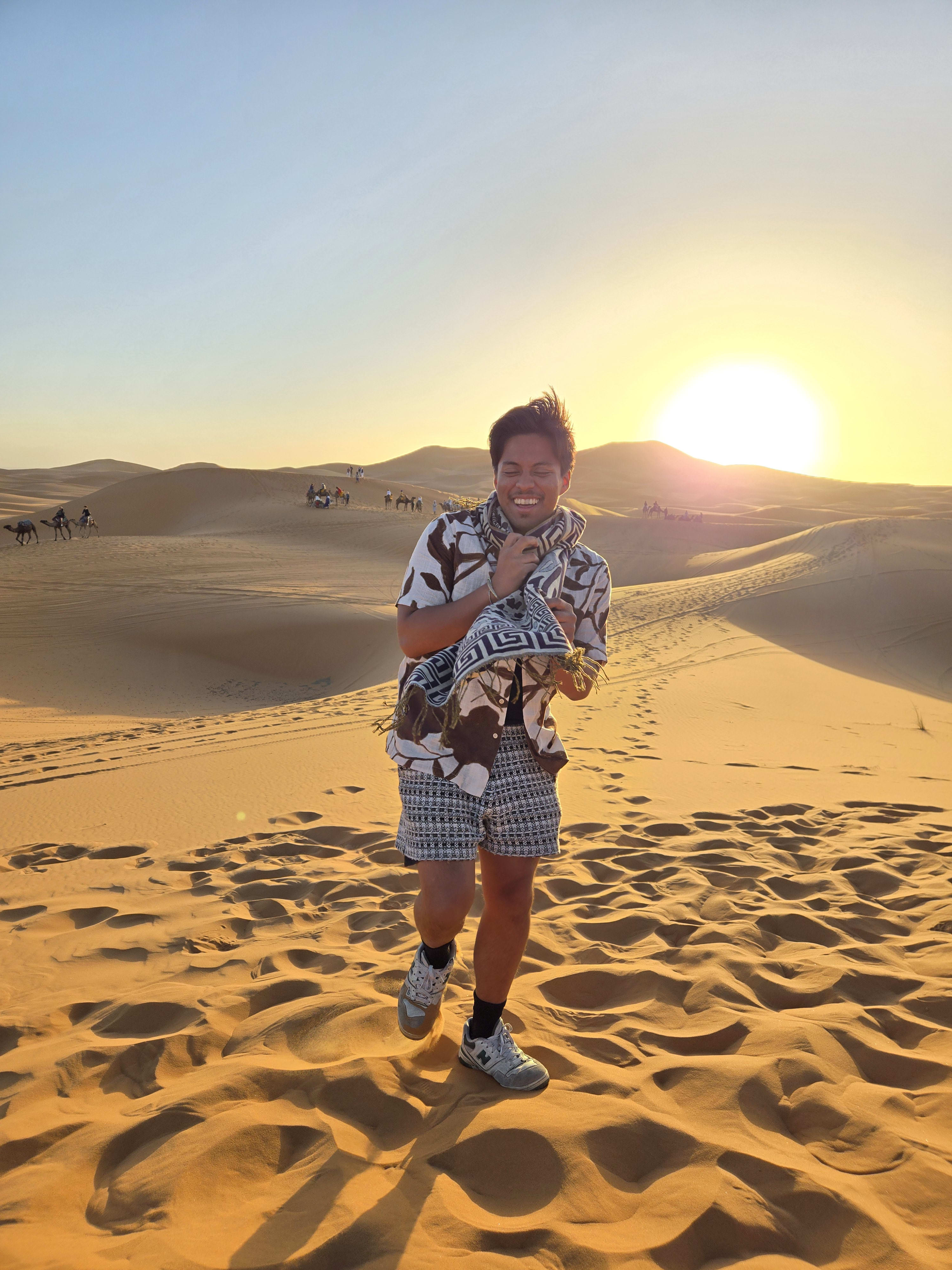 UC Irvine student, John, smiles while standing on a sand dune in the desert with the sun setting behind him.