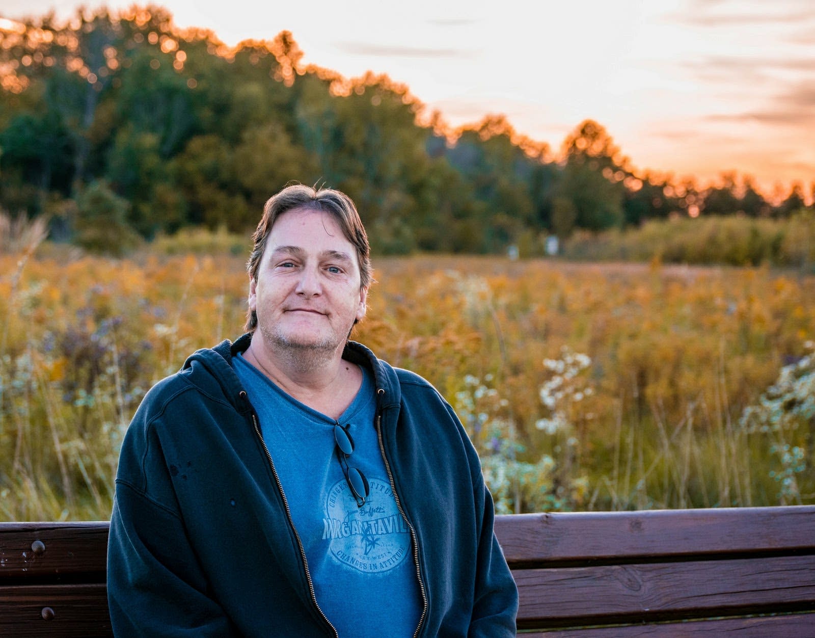 Man smiling while sitting on a bench in field where the sun is setting