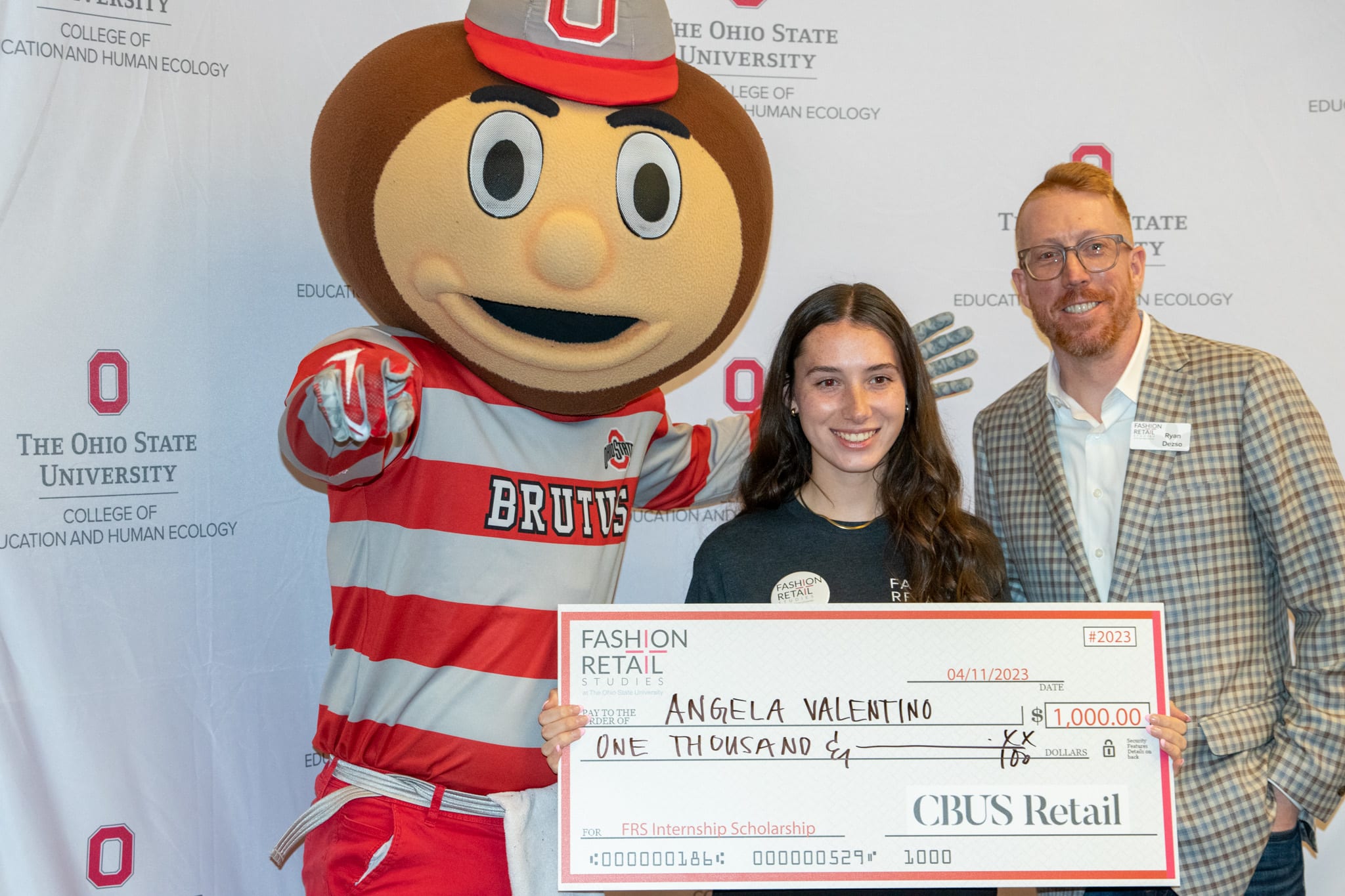 Brutus Buckeye pointing at the camera standing next to two individuals holding a check