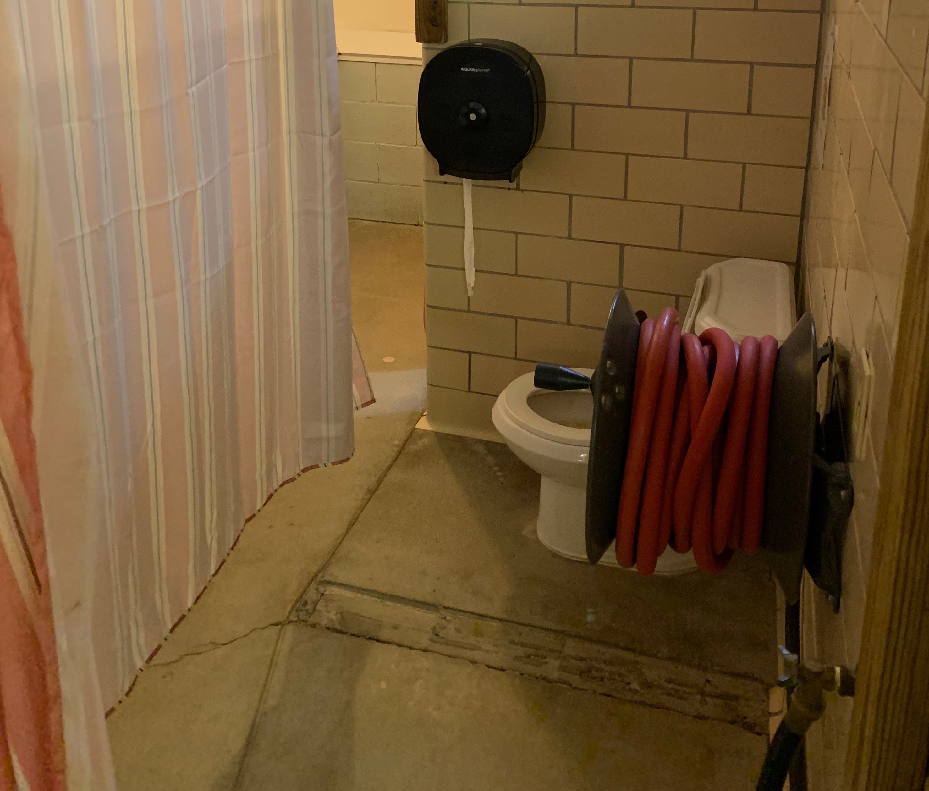 A bathroom stall from camp. A toilet, hose, and a curtain are within this stall
