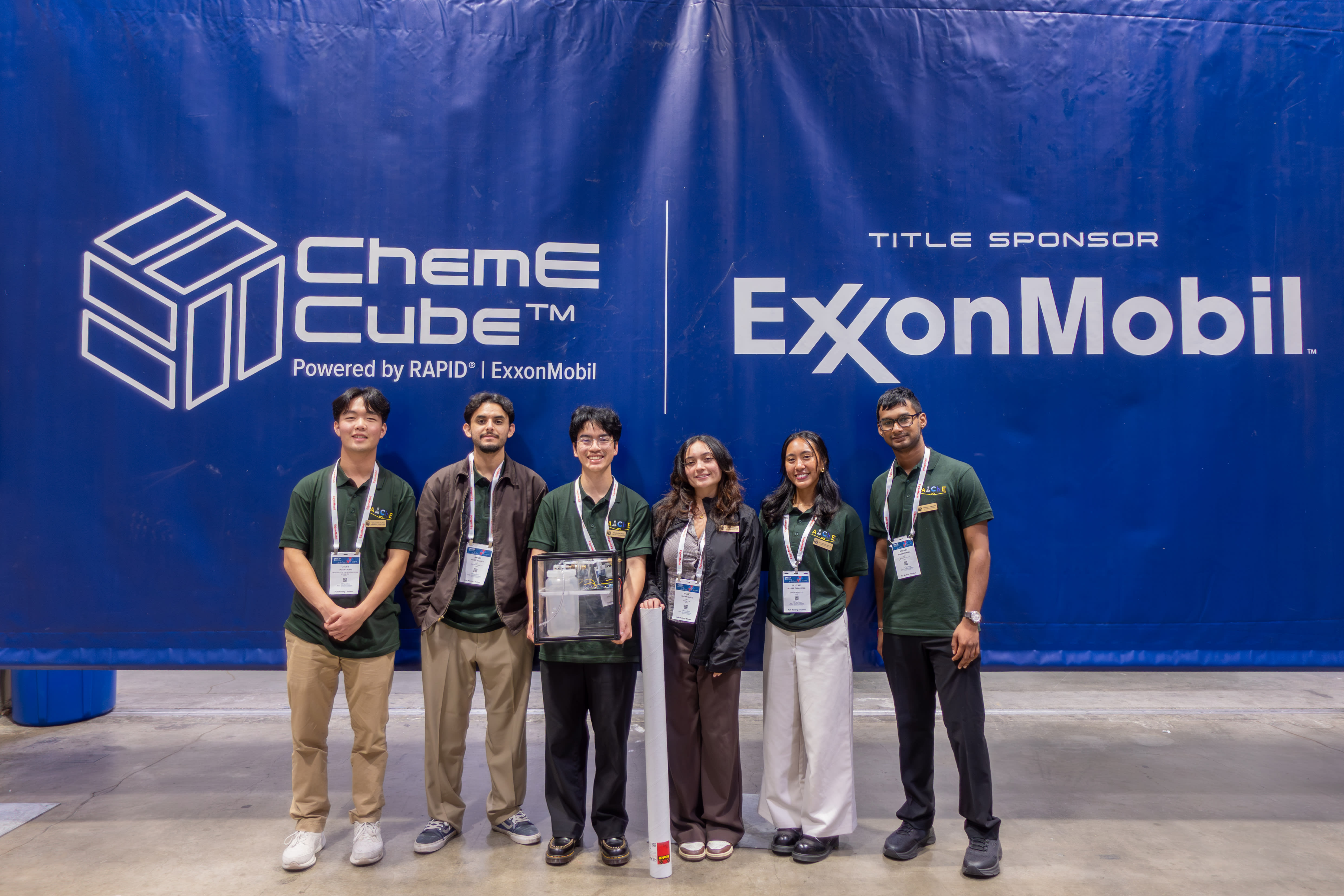 Six member of the ChemE Cube team pose in front of a giant blue banner featuring the ChemE Cube and Exon Mobil logos. All members are wearing matching green T-shirts.