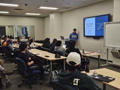 A presenter in a blue "UCI" hoodie stands at the front of a crowded classroom with a presentation on a projector screen.