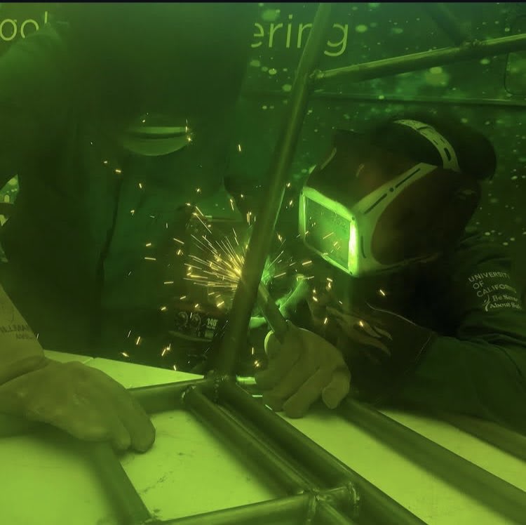 Two students welding in a shop setting with welding masks. The image has a green heugh.