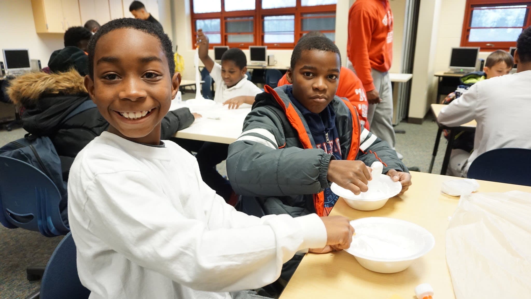 Two young students sit at a table with white Styrofoam bowls on the table 