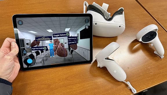 Person holding an iPad in their left hand. iPad is displaying a virtual reality simulation of a human heart. There is a Meta Oculus headset and handset sitting on the a brown table to the right of the iPad.