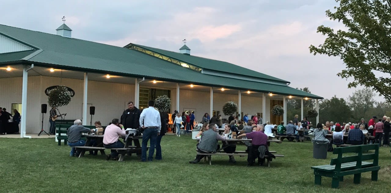 People standing and sitting at picnic tables outside by a barn