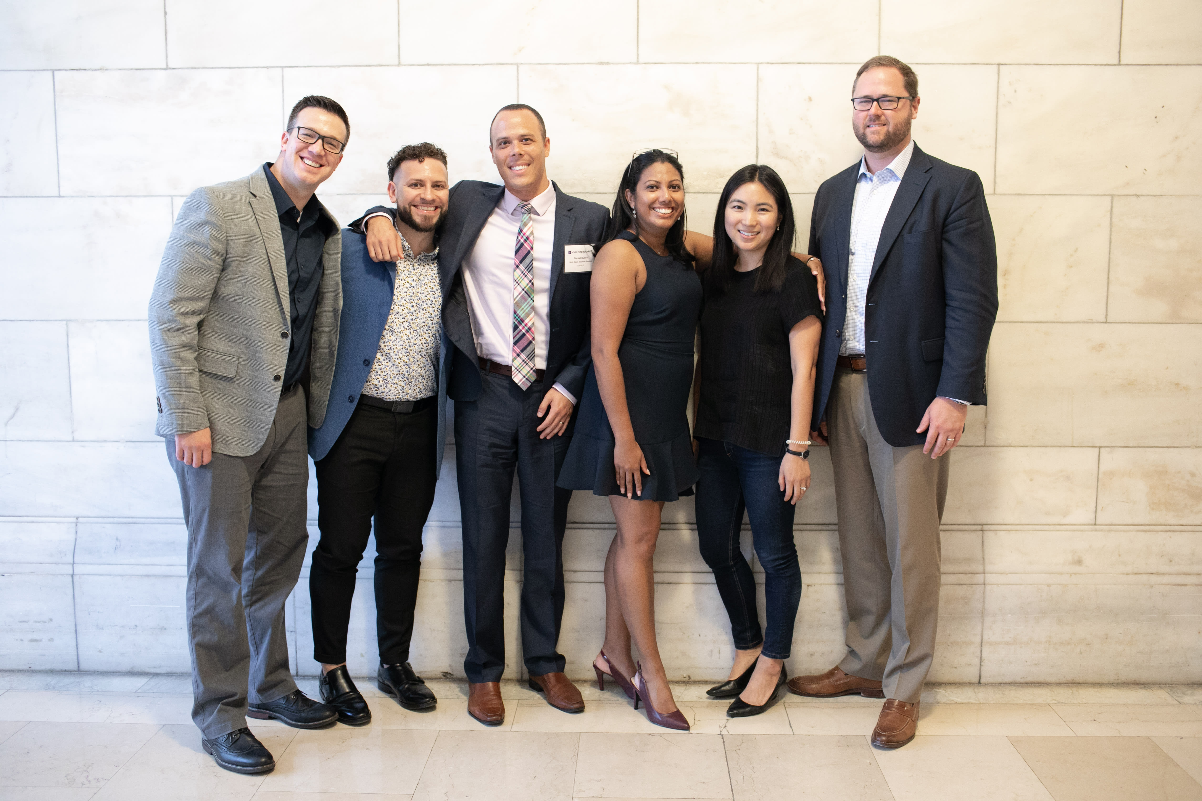 6 people (alumni from cohort 1) gathering for a picture in front of a white tile wall, ready to welcome incoming students.