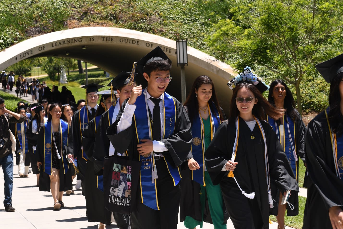 A group of UCI students in graduation gowns walking underneath the PBK bridge at UC Irvine incelebration of the PBK 50th Anniversary at UCI in 2024.