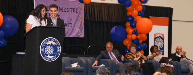 2 students standing at a podium with balloons around them congratulating other graduating students. 