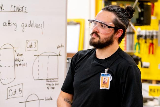 Man with bun, beard, and safety glasses looks at whiteboard with cutting guidelines and diagrams.