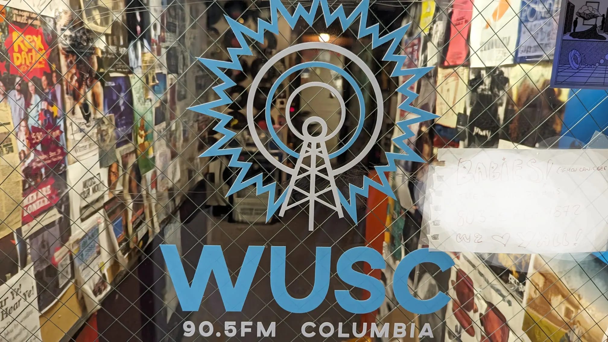 Glass door with WUSC logo and “90.5FM Columbia” text; hallway inside is lined with posters and photos.