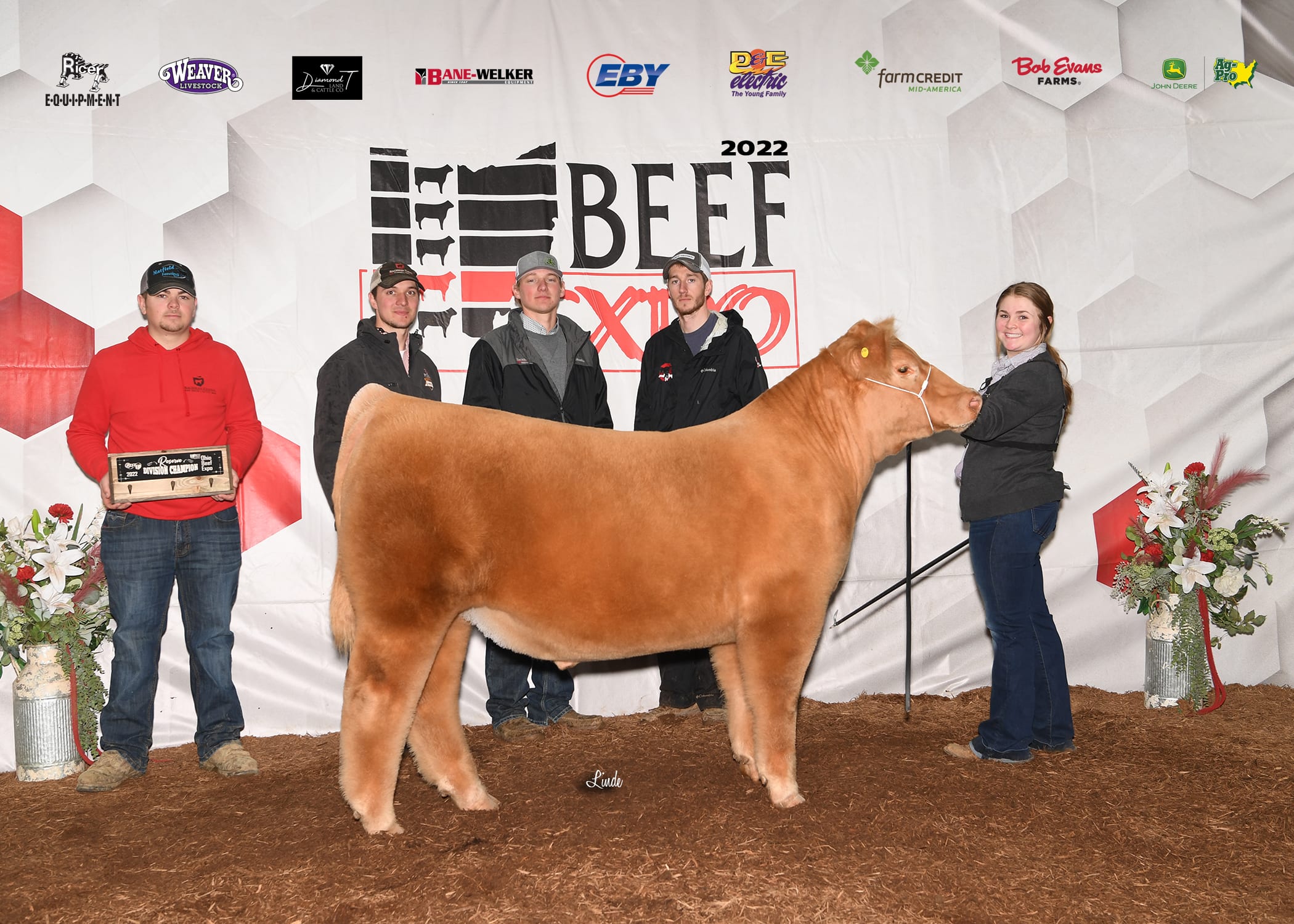 A girl holds her tan cow before the Beef Expo background. Four men stand with her, one holding a multicolored sign