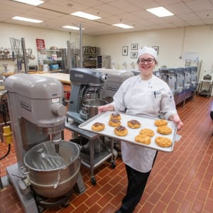 A smiling culinary student in a white chef’s uniform and hat proudly holds a tray of freshly baked pastries in a professional kitchen. Large industrial mixers and baking equipment are visible in the background