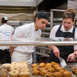 A culinary instructor in a striped apron guides a student in a white chef’s coat and black hat through a pastry display in a professional kitchen. They examine an assortment of freshly baked croissants and bagels on a metal rack.