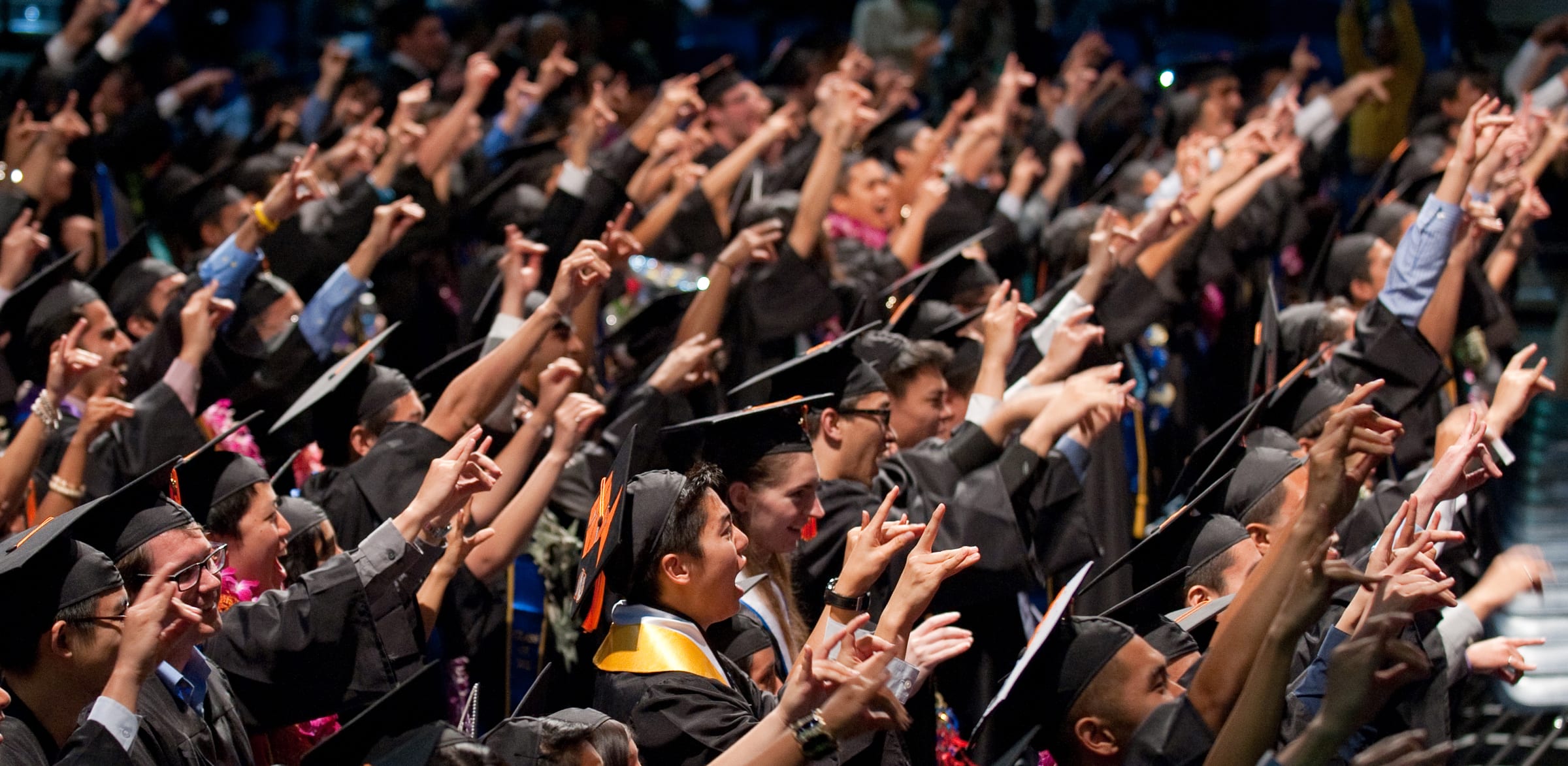 UC Irvine students in caps and gowns seated at commencement, holding up their zots. 