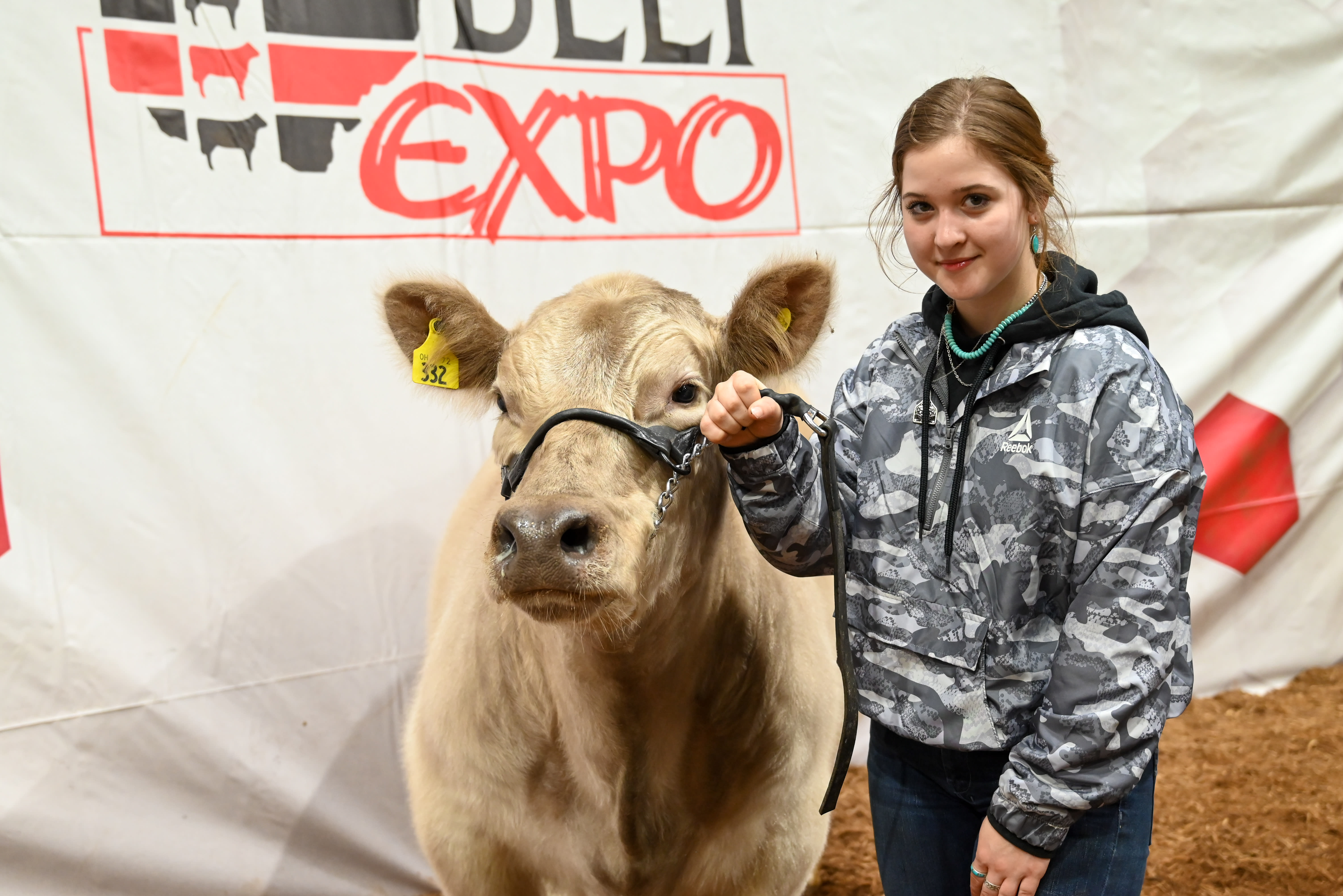 A girl stands to the left of a white steer, holding it close with a rope. They both stand before the Beef Expo backdrop