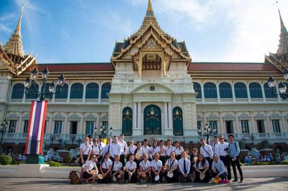 A large group of students, all wearing white tops, pose in front of the Grand Palace in Bangkok, Thailand