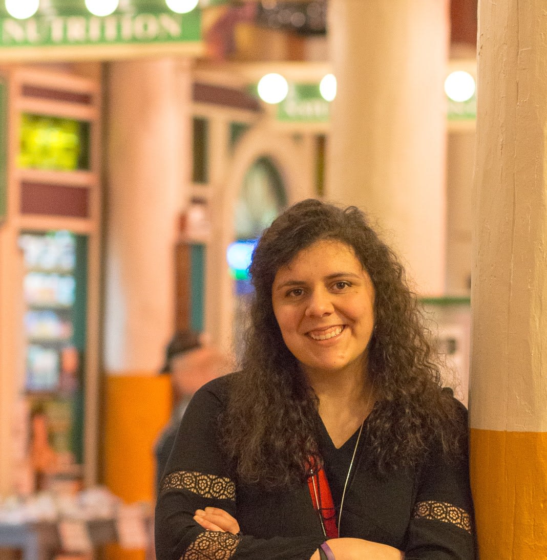 Close up of Lindsay Little, a woman with long curly hair, wearing conference her lanyard and leaning against a pillar.