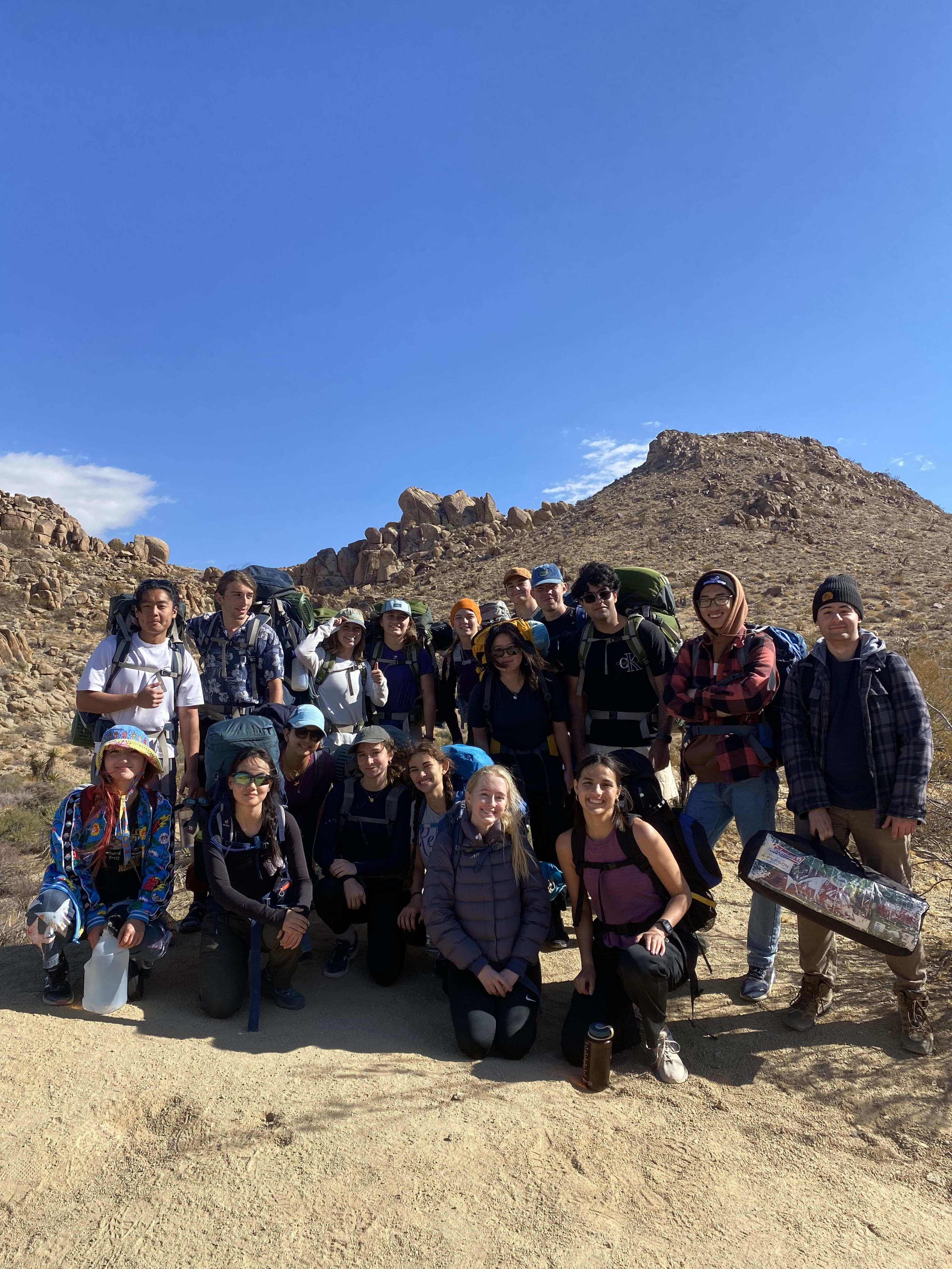 Group of college students stand with backpacking backpacks on and look at the camera. Desert hills are behind them.