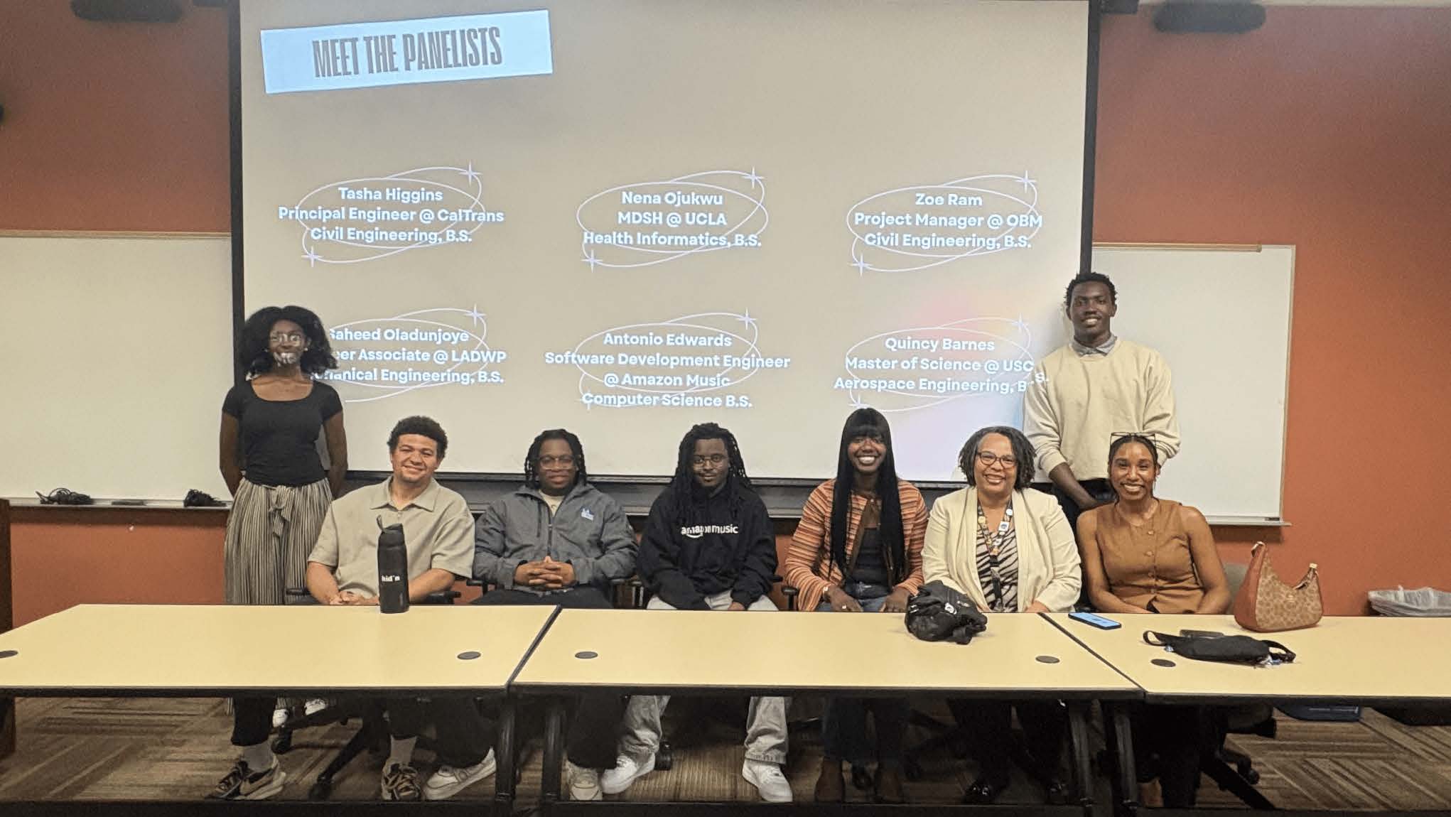Eight alumni sit or stand behind a long table in front of a screen that reads, "Meet the panelists."