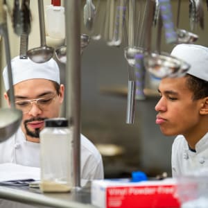 Two culinary students in white chef uniforms and hats focus on a recipe in a professional kitchen. One student, wearing glasses, reads from a sheet of paper, while the other listens attentively. Hanging ladles and whisks frame the scene.